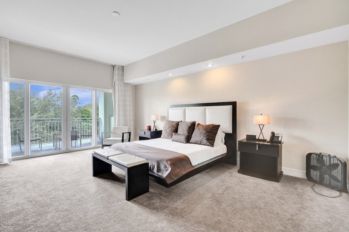 The master bedroom features a spacious layout with a king-size bed adorned with various throw pillows. Natural light floods the room through large sliding glass doors that open to a private veranda, while a bedside lamp and accent chair add functionality.