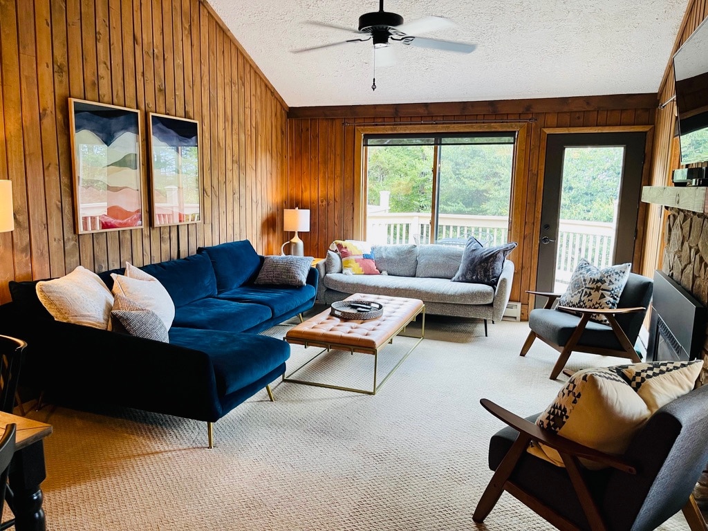 A cozy living area features a blue sectional sofa and a light gray couch, complemented by wooden paneling on the walls. Natural light enters through large windows, illuminating a wooden coffee table and various decorative cushions. A lounge chair and a ceiling fan add to the comfortable ambiance.