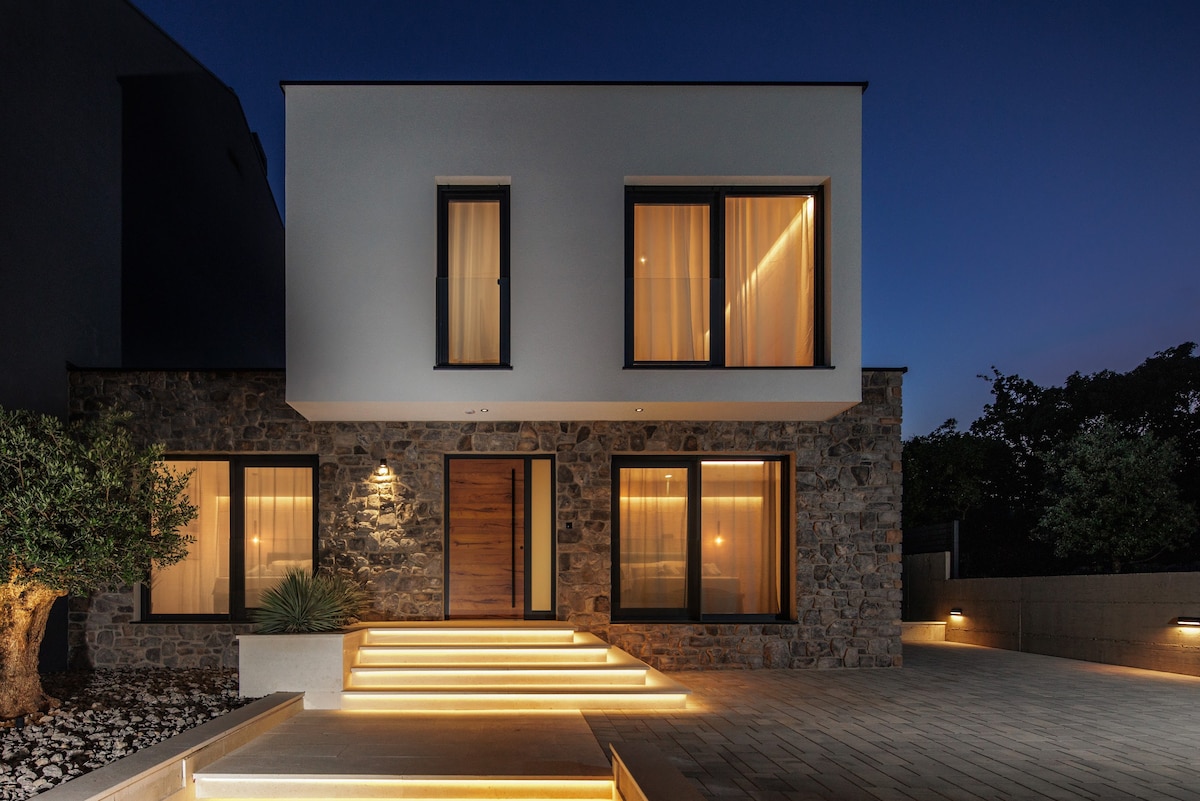 The exterior of the modern villa is presented at dusk, featuring large glass windows and a bold wooden door. Soft lighting highlights the stone facade and illuminated steps leading to the entrance. Lush greenery surrounds the property, adding a natural touch to the contemporary design.