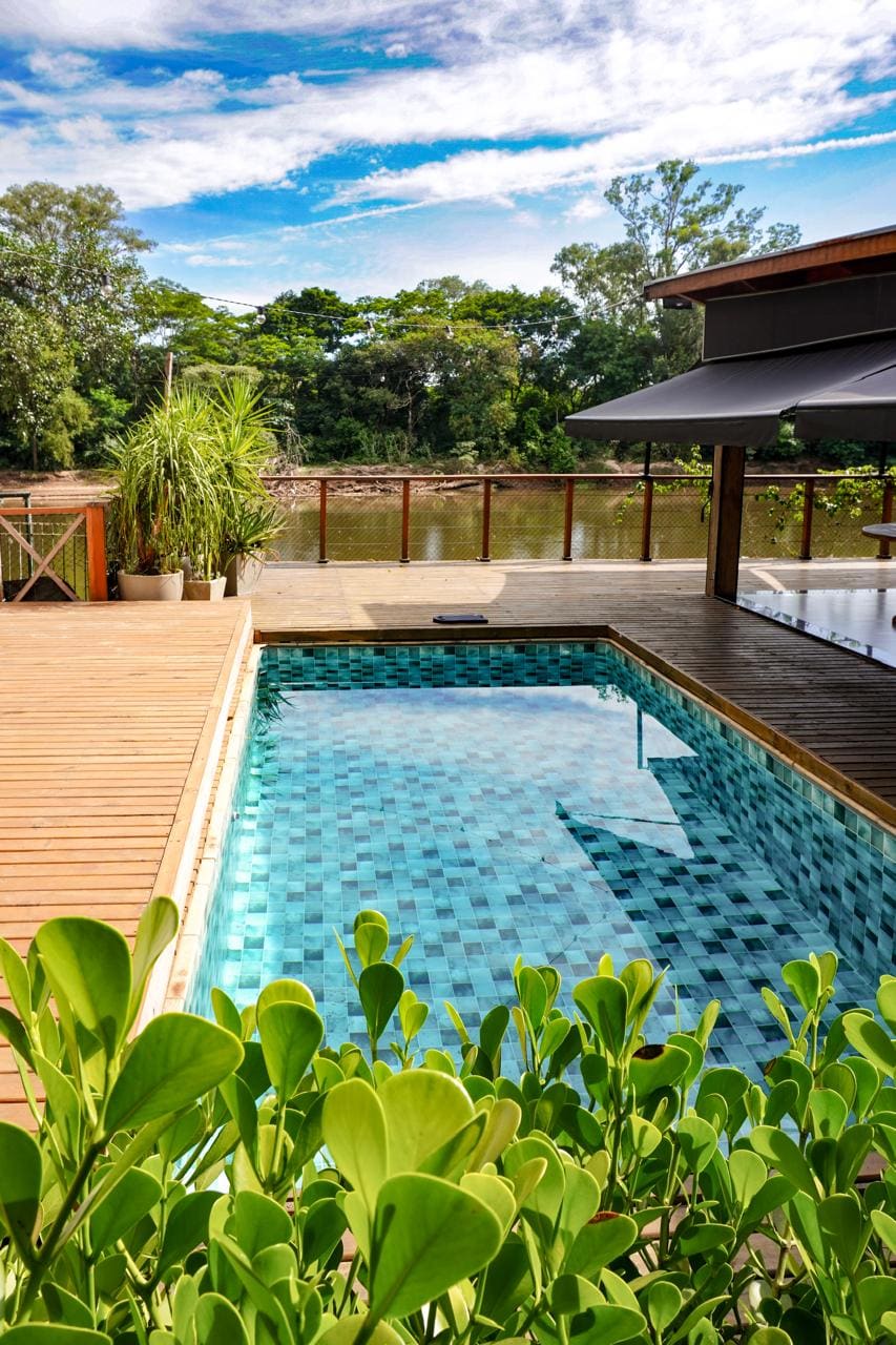 A refreshing pool with a tiled surface is framed by wooden decking, creating an inviting space for relaxation. Lush greenery lines the foreground, while the serene river and trees can be seen in the background under a clear sky.