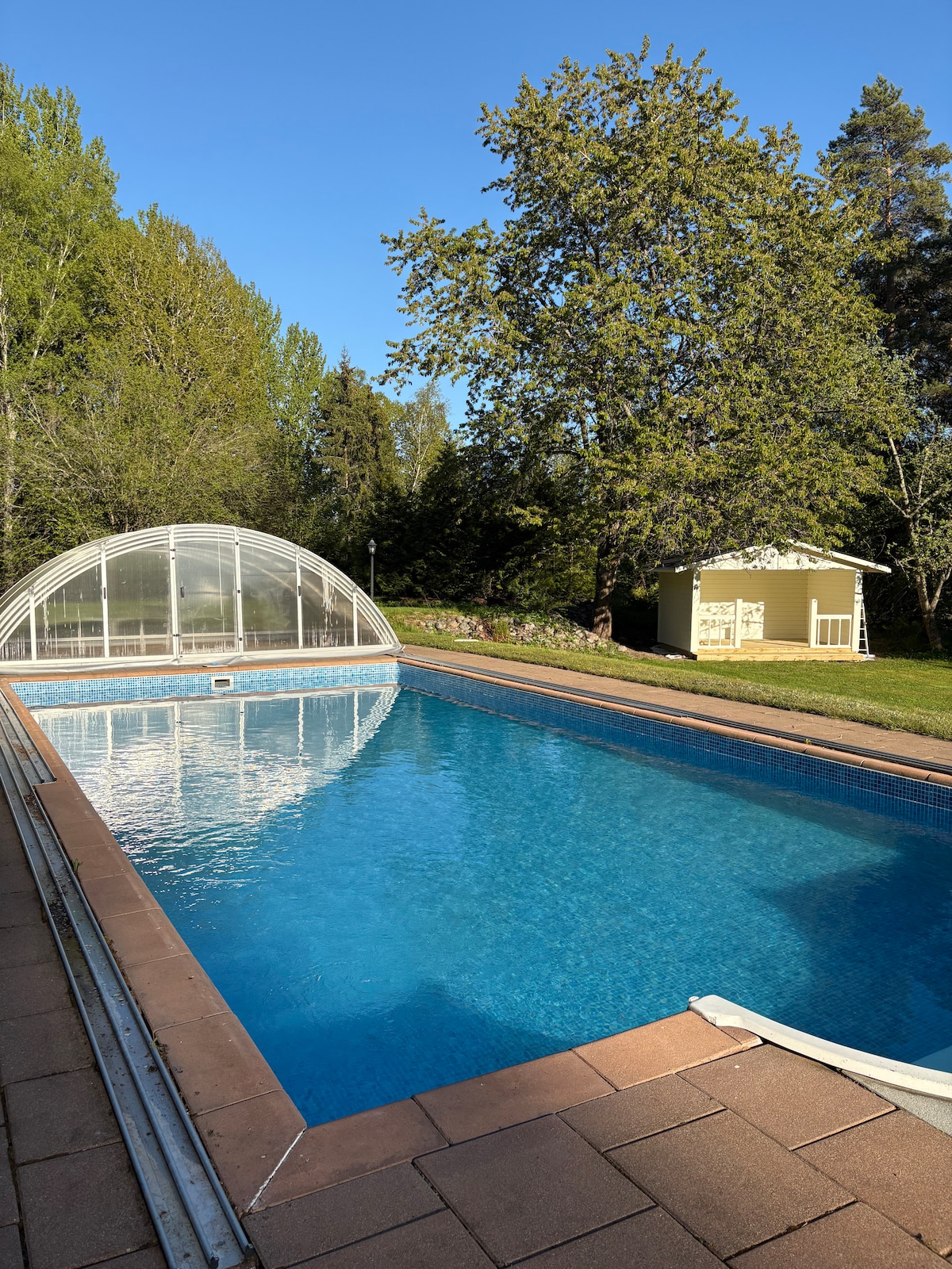 A large outdoor swimming pool is framed by a paved area, with clear water reflecting the blue sky. A protective cover is visible over part of the pool. Lush greenery surrounds the space, including tall trees and a small storage shed in the background.