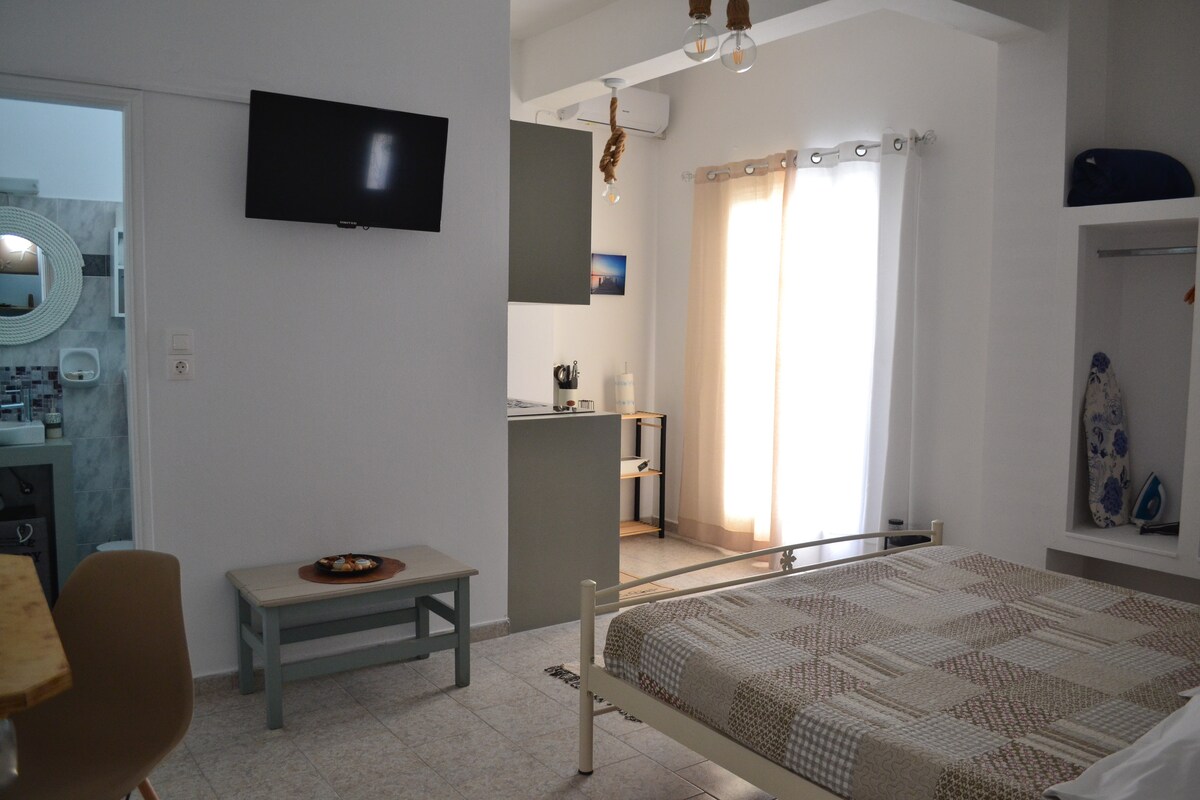 The interior of the studio is shown with a spacious bed at the forefront, surrounded by light-colored walls. A kitchenette is visible in the background, along with a small dining area. Natural light streams in from the window, enhancing the open and airy feel of the space.