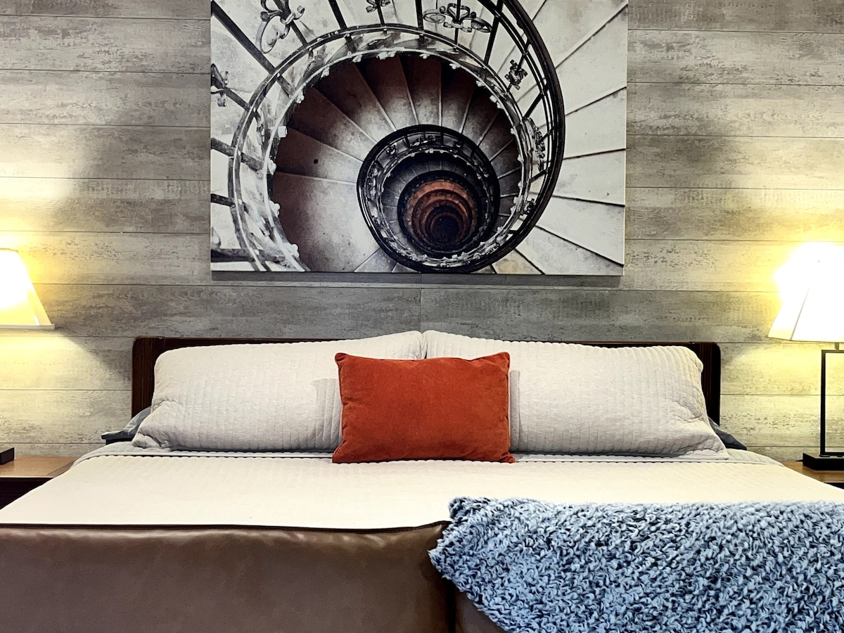A spacious bed is centered in the room, complemented by a decorative orange pillow and a cozy blue throw. A striking black and white spiral staircase photograph adorns the wall above the bed. Soft lighting is provided by bedside lamps on each side.