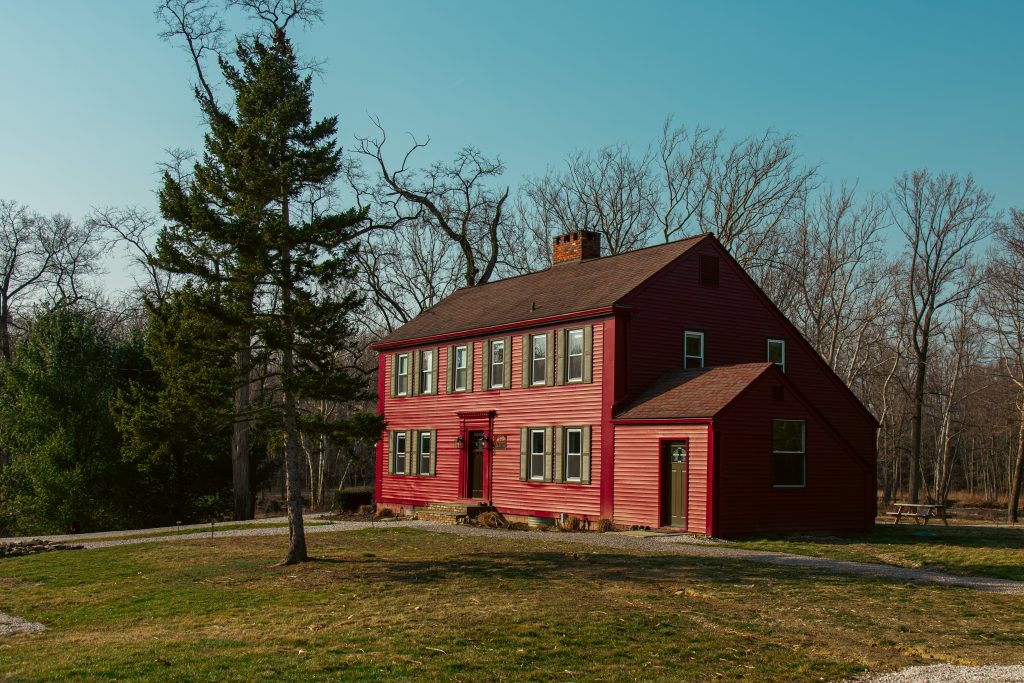 A historic red saltbox lodge is showcased amidst a natural setting. The structure features multiple windows and a prominent chimney. Surrounding grassy areas and trees provide a serene backdrop against the clear blue sky.