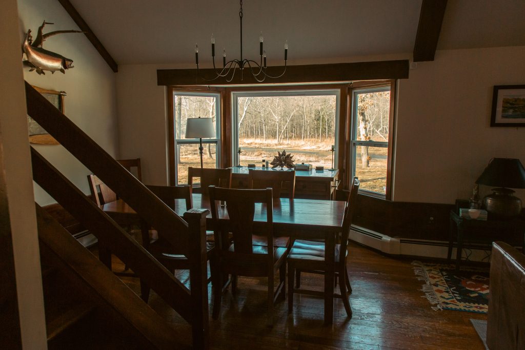A dining area features a large wooden table surrounded by several chairs, with natural light streaming in from large windows. A chandelier hangs above the table, while decorative elements and a sideboard enhance the space. Outdoor scenery is visible through the windows.