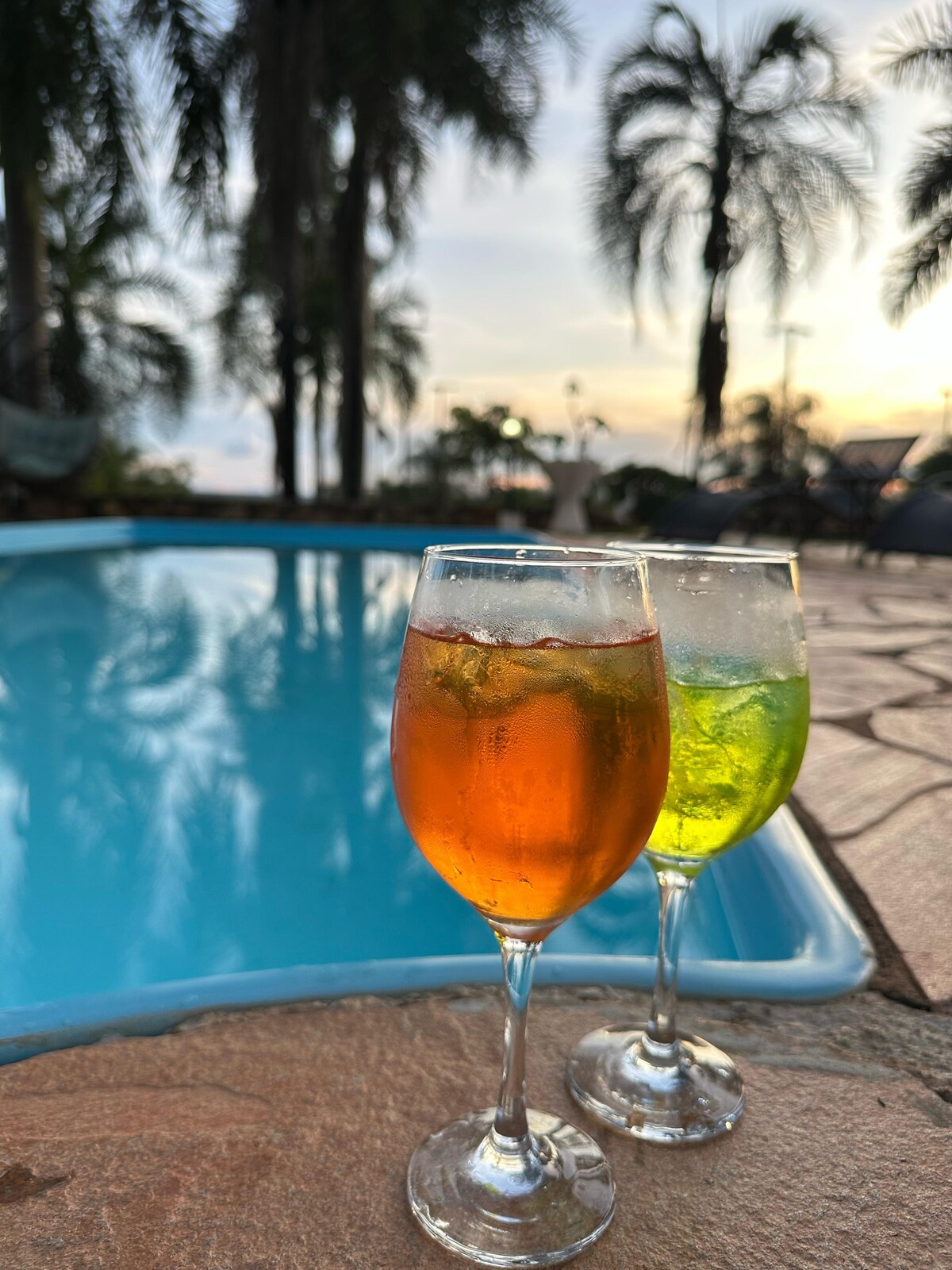 Two elegant glasses sit on a stone surface beside a clear pool, with one containing a vibrant orange beverage and the other a refreshing green drink. Lush palm trees can be seen in the background, framing a serene sunset sky.