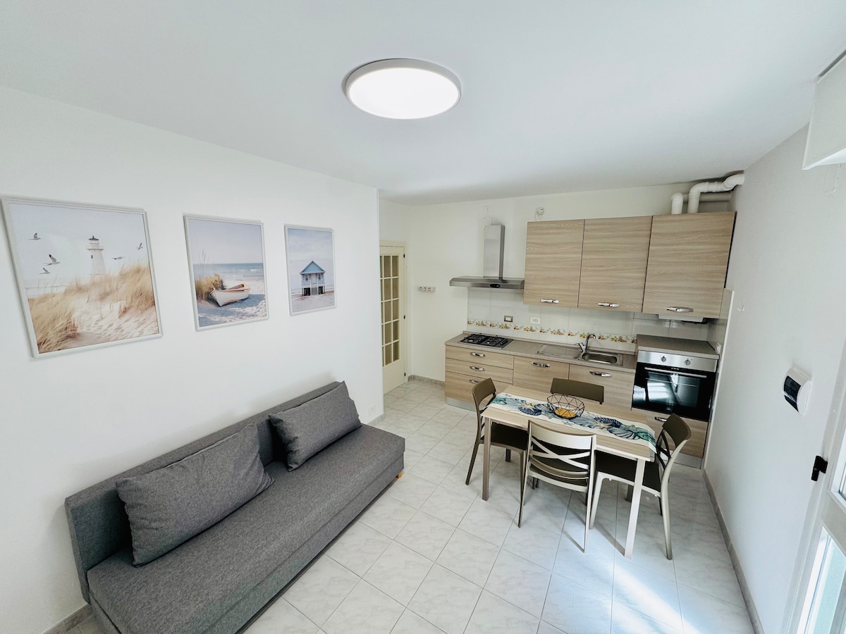 A spacious living area is featured, showcasing a gray sofa facing a dining table set for four. The kitchen area includes modern cabinets and appliances. Three framed images depicting coastal scenes adorn the wall, and natural light fills the room through a nearby window.