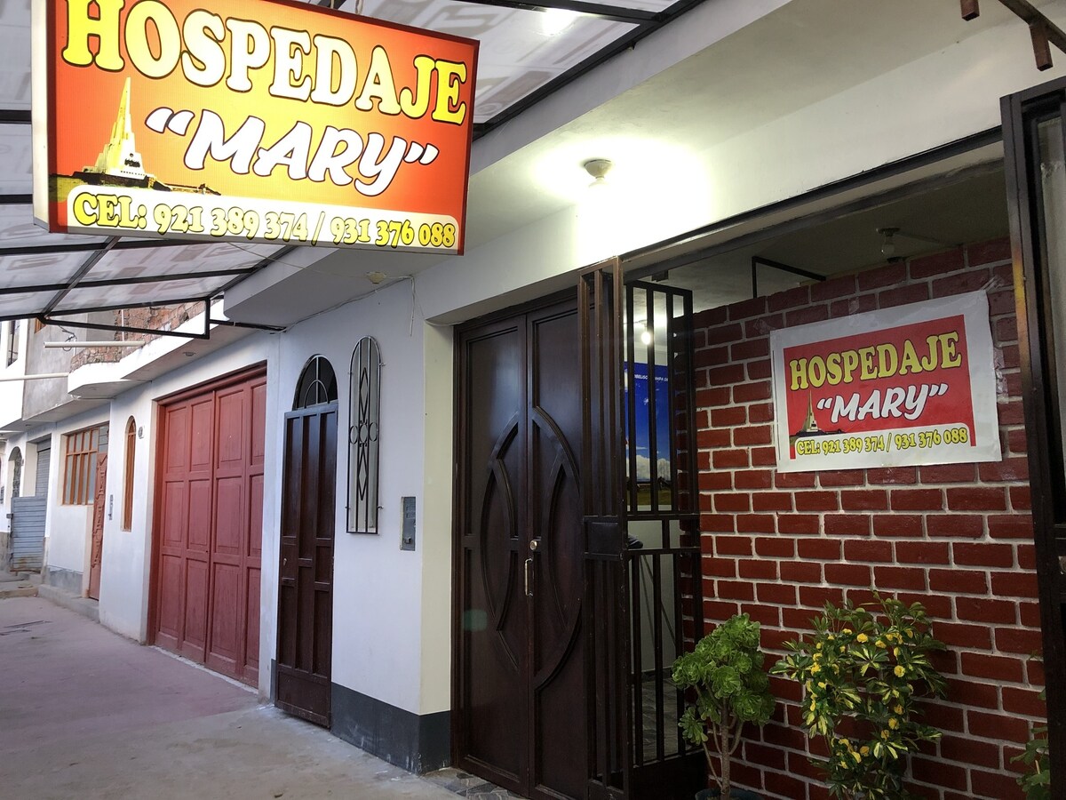 The entrance of Hospedaje Mary is framed by contrasting brick and wooden elements. A bright sign displays the name prominently above, while a glass door allows visibility into the welcoming interior. The pathway is lined by simple decorative features, creating an inviting appearance.