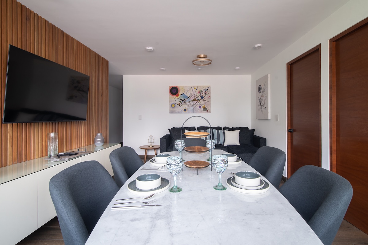 An elegant dining area features a modern oval table set with six plates and glassware. The space is complemented by a sleek sideboard and a wall-mounted TV. Artwork adds a touch of sophistication, while large windows provide natural light.