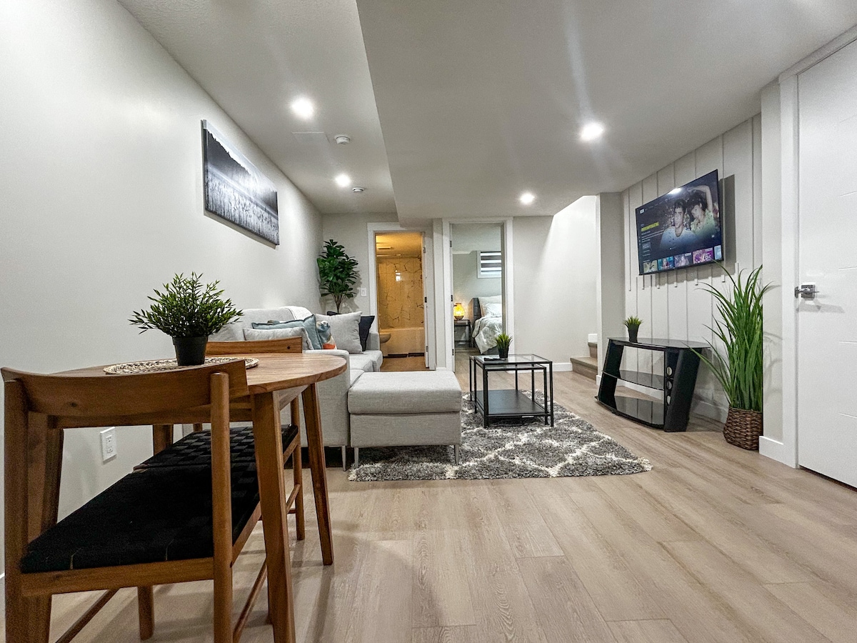 A modern living area features a cozy seating arrangement with a sectional sofa and an accent chair. A small dining table with two chairs is present, alongside a coffee table. Green plants and a media console with a Smart TV enhance the space, surrounded by light wood flooring.