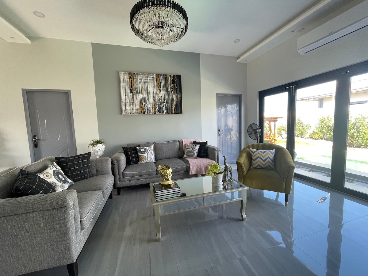 A modern living room is presented, featuring a gray sectional sofa and an accent chair in vibrant yellow. A glass coffee table is centered on glossy tiled flooring, with decorative items atop it. Large glass doors provide views of the garden, enhancing the natural light.