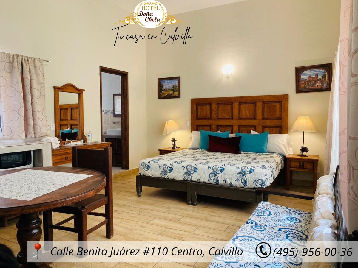 A spacious suite features a large bed with decorative pillows and a colorful bedspread. A wooden dresser and a round dining table with chairs are present. Natural light enters through a window with sheer curtains, creating a bright atmosphere. Artwork adorns the walls.