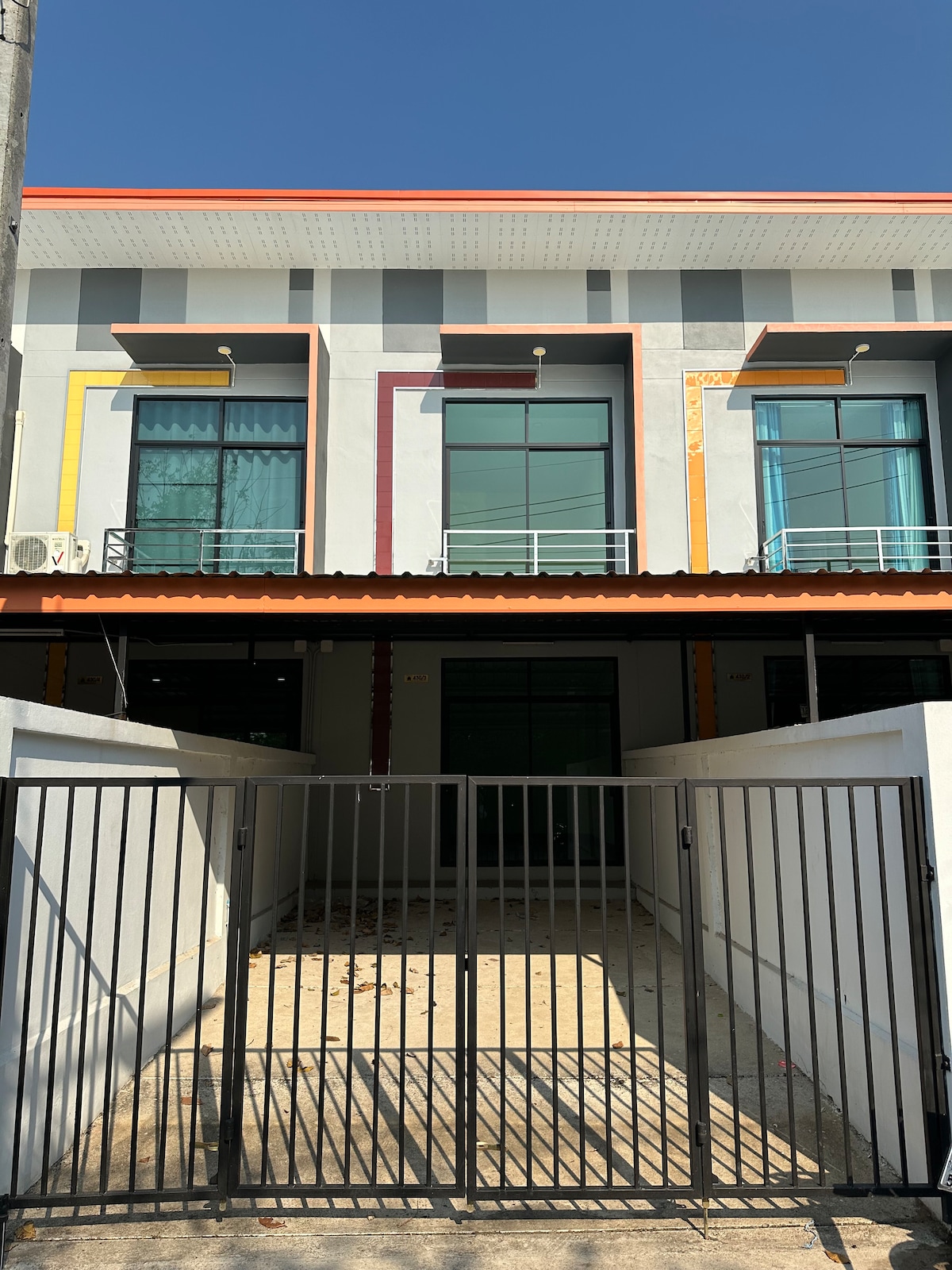 A modern two-story building is framed by a sleek black gate. Large glass windows, adorned with colorful accents, invite natural light into the space. The entrance is set against a clear blue sky, emphasizing the structure's contemporary design.