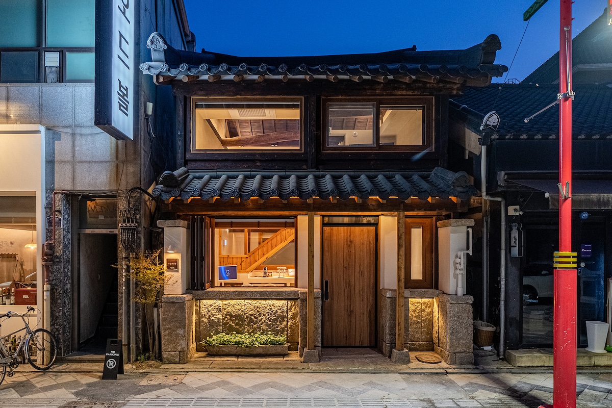 The two-story traditional house is framed by large glass windows, reflecting the evening light. Natural wooden details enhance the façade, while warm light glows from the entrance, inviting guests in. The surrounding area features a cobblestone path and nearby modern buildings.