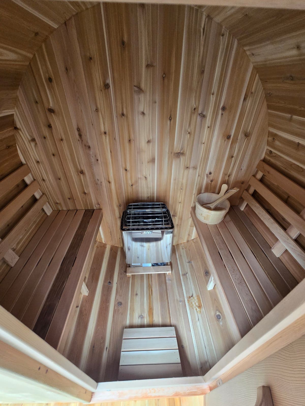 A wooden sauna interior showcases warm cedar paneling, creating a cozy atmosphere. A small stove is present for heating, with wooden benches lining the sides. A wooden bucket and ladle are neatly arranged, providing amenities for sauna use.