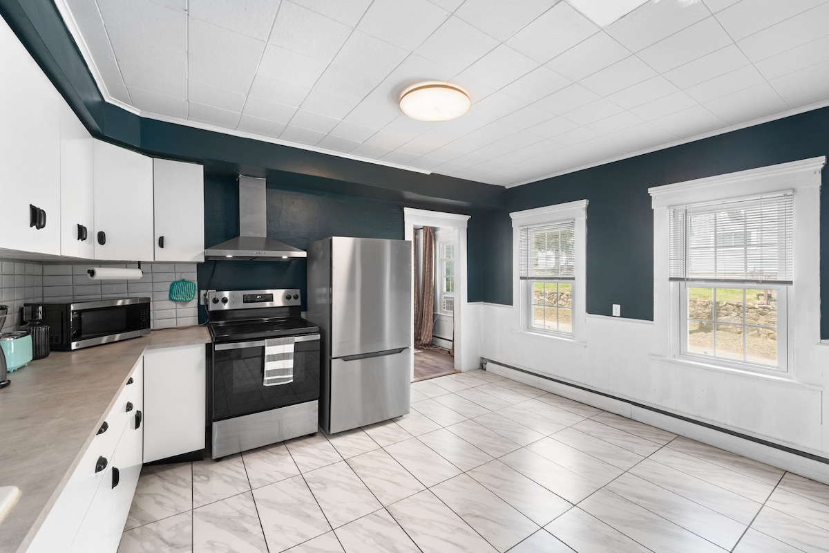 The kitchen features a modern stainless steel refrigerator and stove, complemented by white cabinetry and a dark accent wall. A spacious layout is highlighted by a tiled floor, providing ample room for cooking and dining. Natural light enters through the windows, enhancing the fresh atmosphere.
