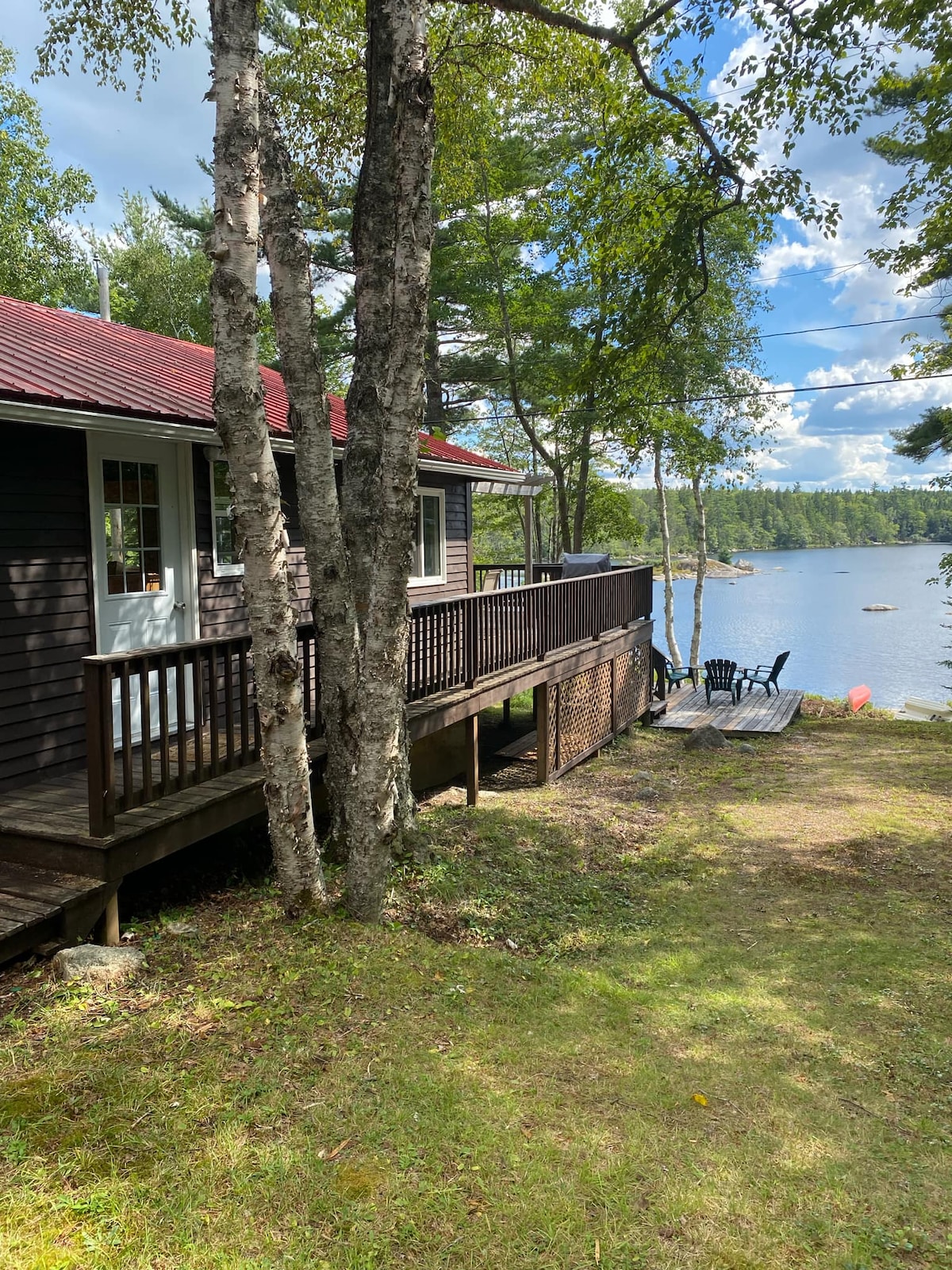 A rustic cabin is seen by the lakeside, surrounded by trees. A wooden deck with seating overlooks the water, allowing for easy access. The setting is tranquil, offering a serene environment for relaxation, while the cabin's exterior reflects natural tones under a clear blue sky.