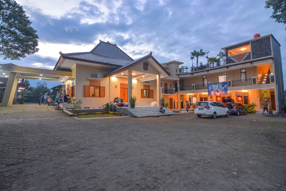 The property features a spacious parking area surrounded by well-maintained grounds. The main building reveals a traditional architectural style with a peaked roof, while a modern structure is visible alongside. Guests can be seen enjoying the outdoor space under a cloudy sky.