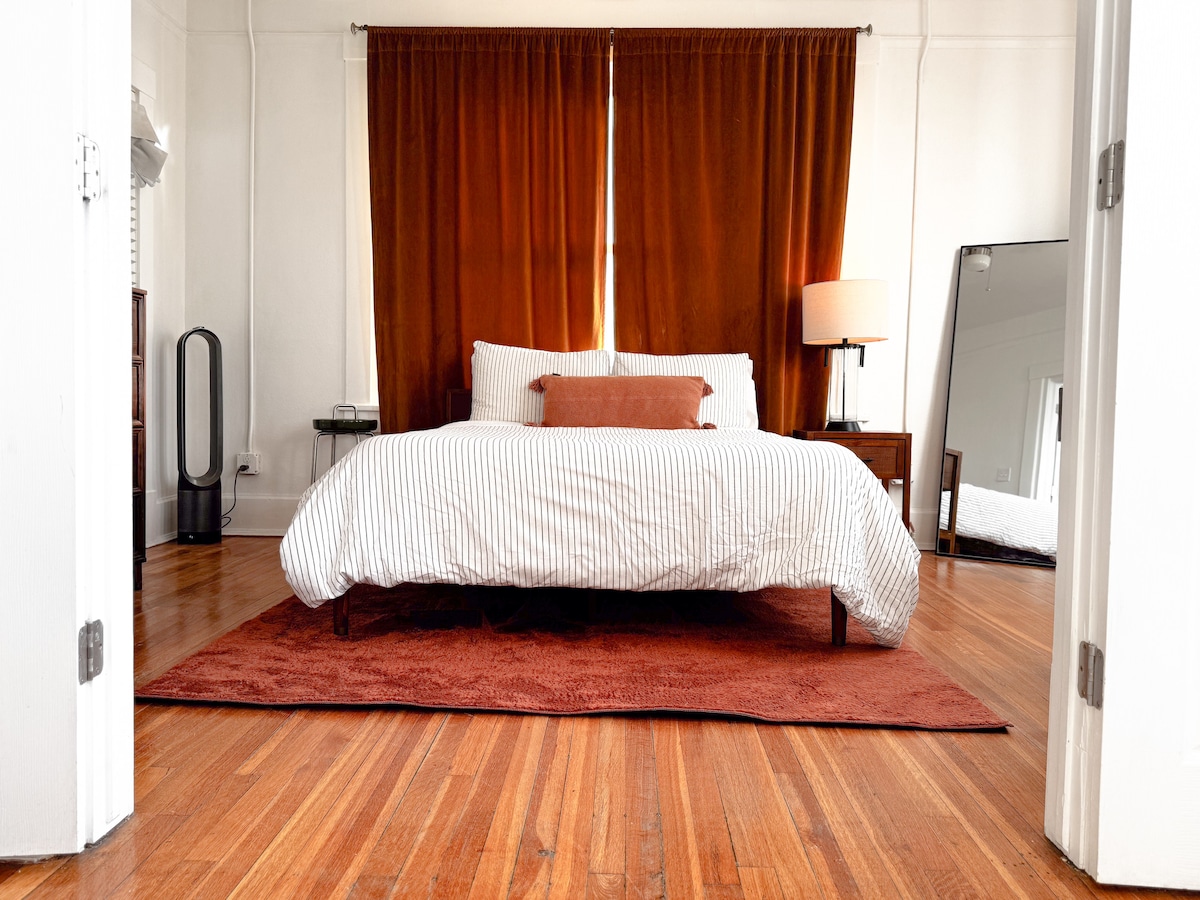 The bedroom features a spacious bed with white and striped bedding, accompanied by an orange accent pillow. Natural light enters through a window, illuminating the wooden floors and warm tones of a large area rug. A full-length mirror and bedside table add functional charm.