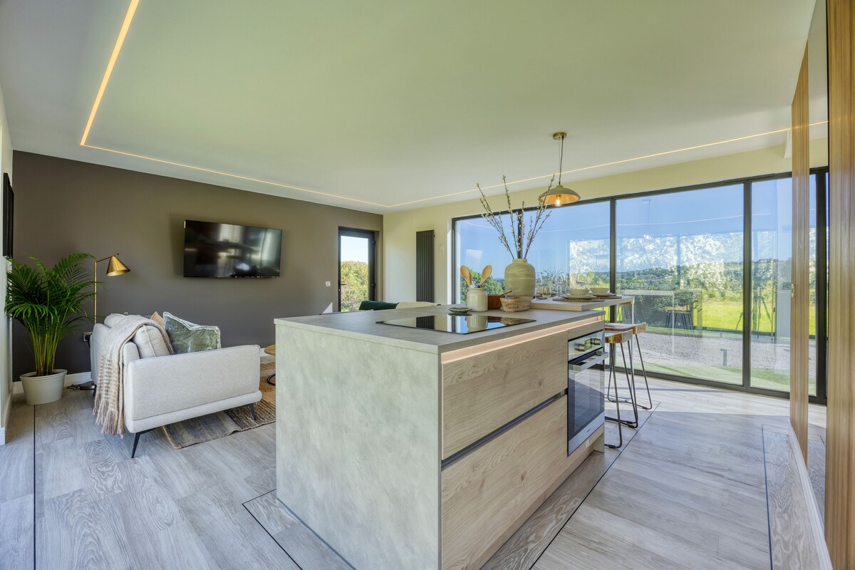 An open-plan living area is showcased, featuring a modern kitchen island with seating. Large glass windows provide panoramic views of the serene landscape, allowing natural light to fill the space. A comfortable sofa and a flat-screen TV are positioned nearby, enhancing the inviting atmosphere.