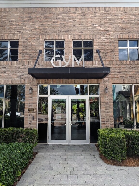The entrance to a gym is framed by two large glass doors, allowing visibility into the interior. Above the doors, the word 'GYM' is displayed prominently. The exterior features a classic brick facade and is surrounded by neatly trimmed shrubs, adding to the welcoming appearance.