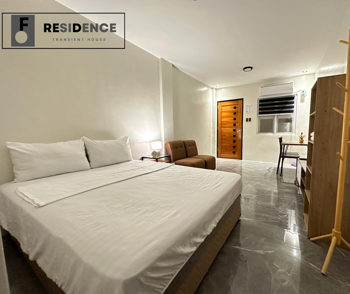A well-lit room features a spacious bed with crisp white linens. A comfortable brown sofa is positioned beside the bed. A small wooden table and chair are placed against the wall, with a window allowing natural light to stream in.