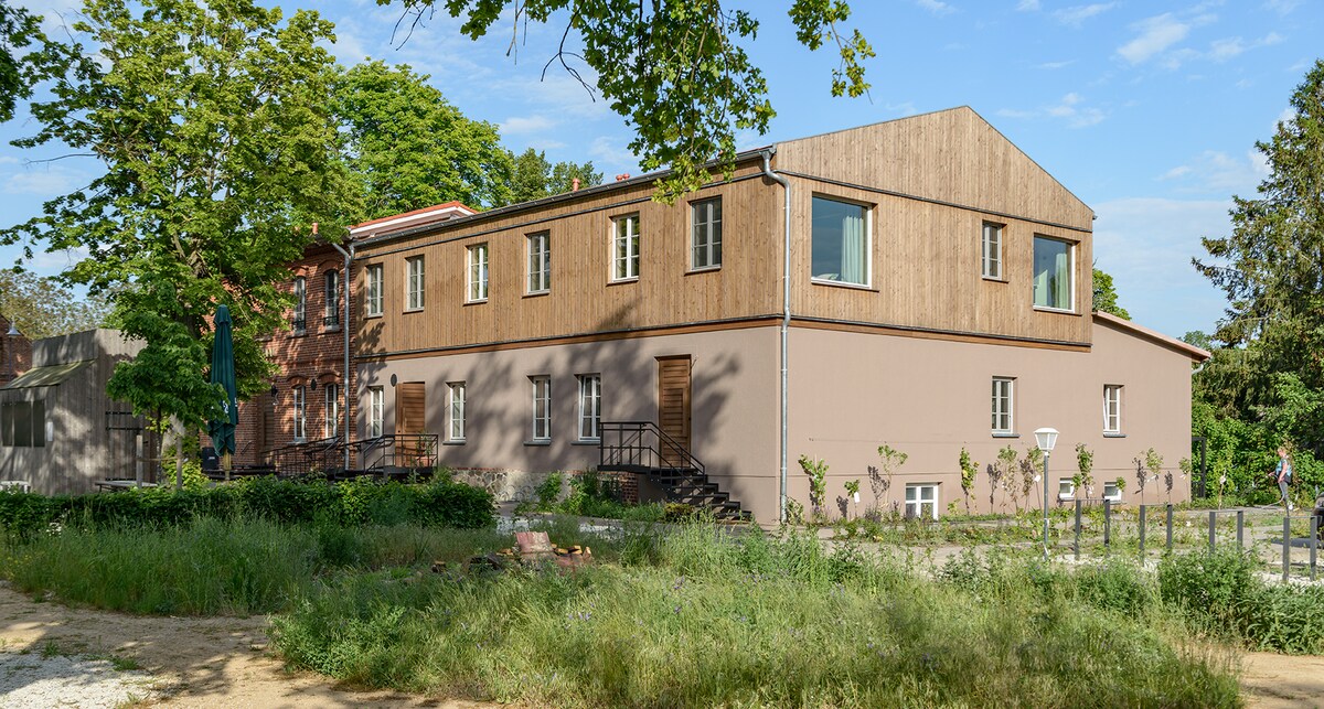The exterior of a two-story building is visible, featuring a combination of wooden and stucco materials. Several windows allow natural light to enter, while steps lead to the main entrance. Lush greenery surrounds the structure, contributing to a tranquil setting.