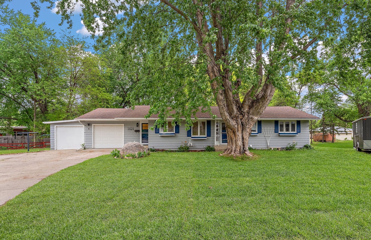 A single-story home is shown, surrounded by lush green grass and mature trees. The exterior features a light-colored facade, complemented by blue window shutters. A long driveway leads to a garage on the left, while a spacious front yard is partially shaded by a large tree.