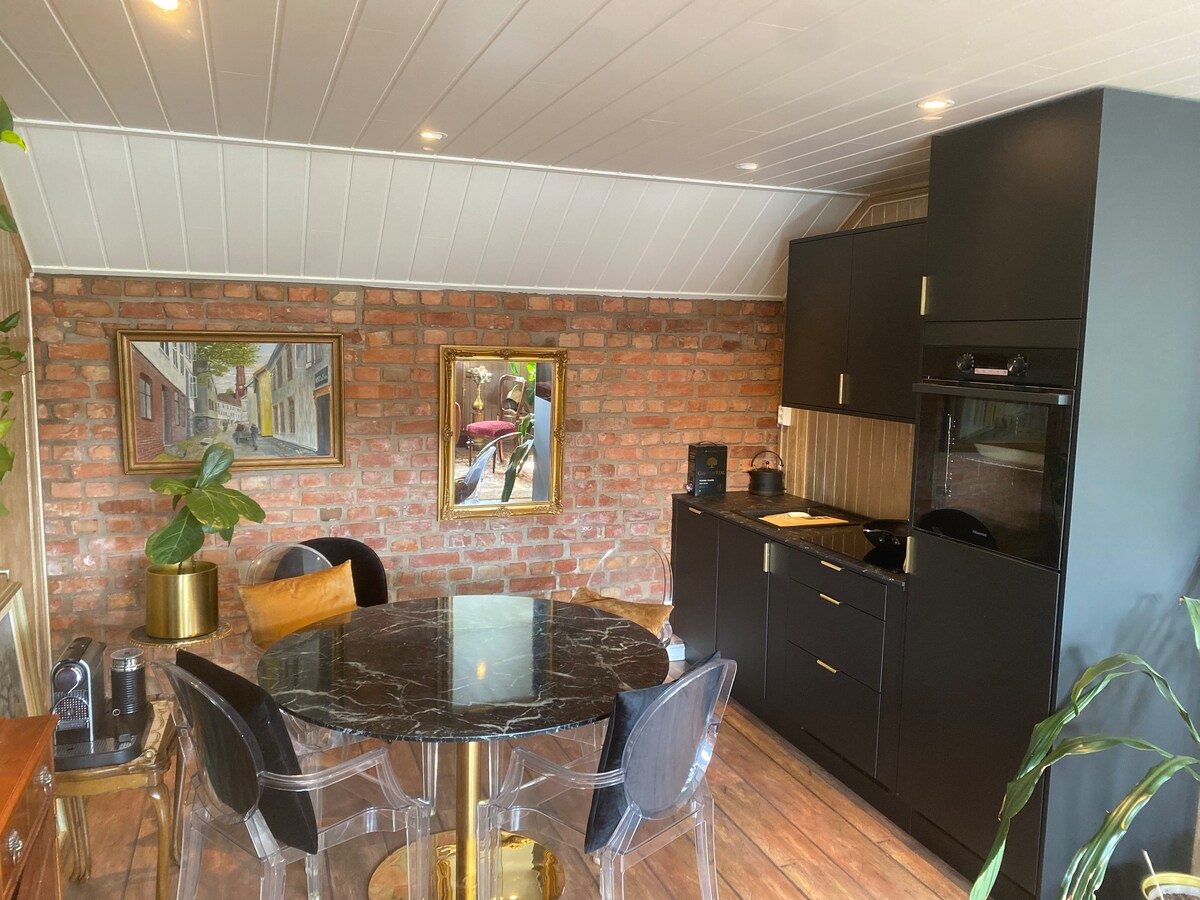 A modern kitchen area is showcased, featuring dark cabinetry and a marble dining table surrounded by black chairs. Exposed brick walls add character, while greenery is present, enhancing the inviting ambiance of the space.