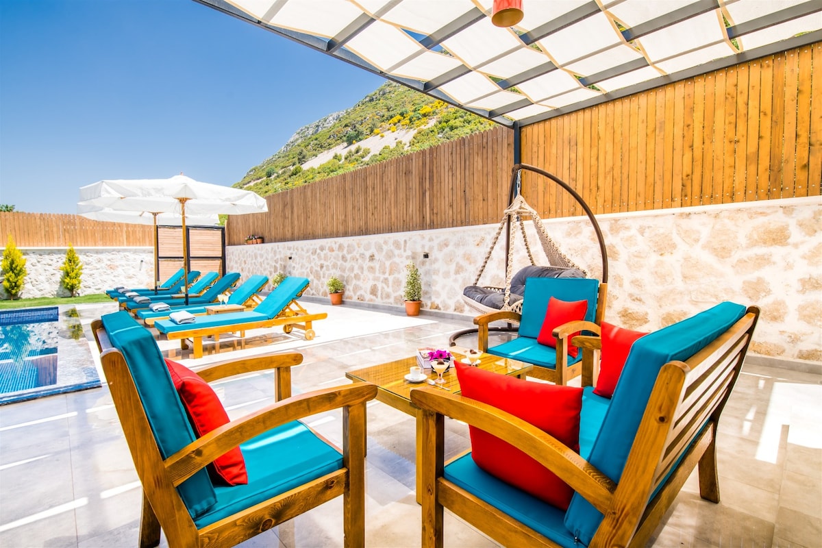An outdoor seating area features comfortable wooden chairs with bright cushions, inviting relaxation. A table sits in the center, adorned with floral arrangements. A hammock chair swings gently nearby, surrounded by sun loungers and vibrant umbrellas, all set against a backdrop of natural greenery and stone walls.