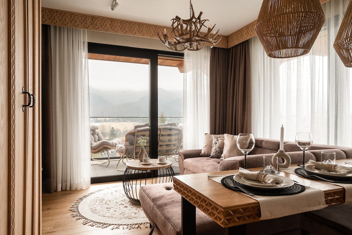 An elegant living area features a spacious seating arrangement with a textured sofa, complemented by a wooden coffee table. Natural light floods the room through expansive glass doors, revealing a scenic balcony overlooking the mountains. Textured light curtains add a soft touch to the ambiance.