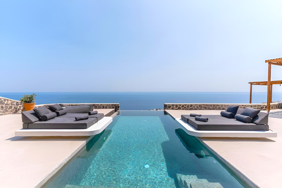 An infinity pool extends into the horizon, bordered by two lounge beds. The calm blue water of the pool reflects the sky, creating a seamless view of the sea. A pergola provides shade to one side, enhancing the serene setting.