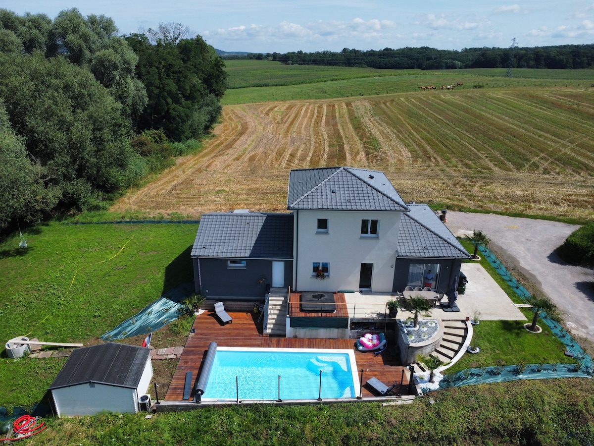 An aerial view captures a modern home set within a lush green landscape. A spacious pool is surrounded by a wooden deck and sun loungers, complemented by a jacuzzi nearby. Fields stretch into the distance, enhancing the tranquility of the setting.