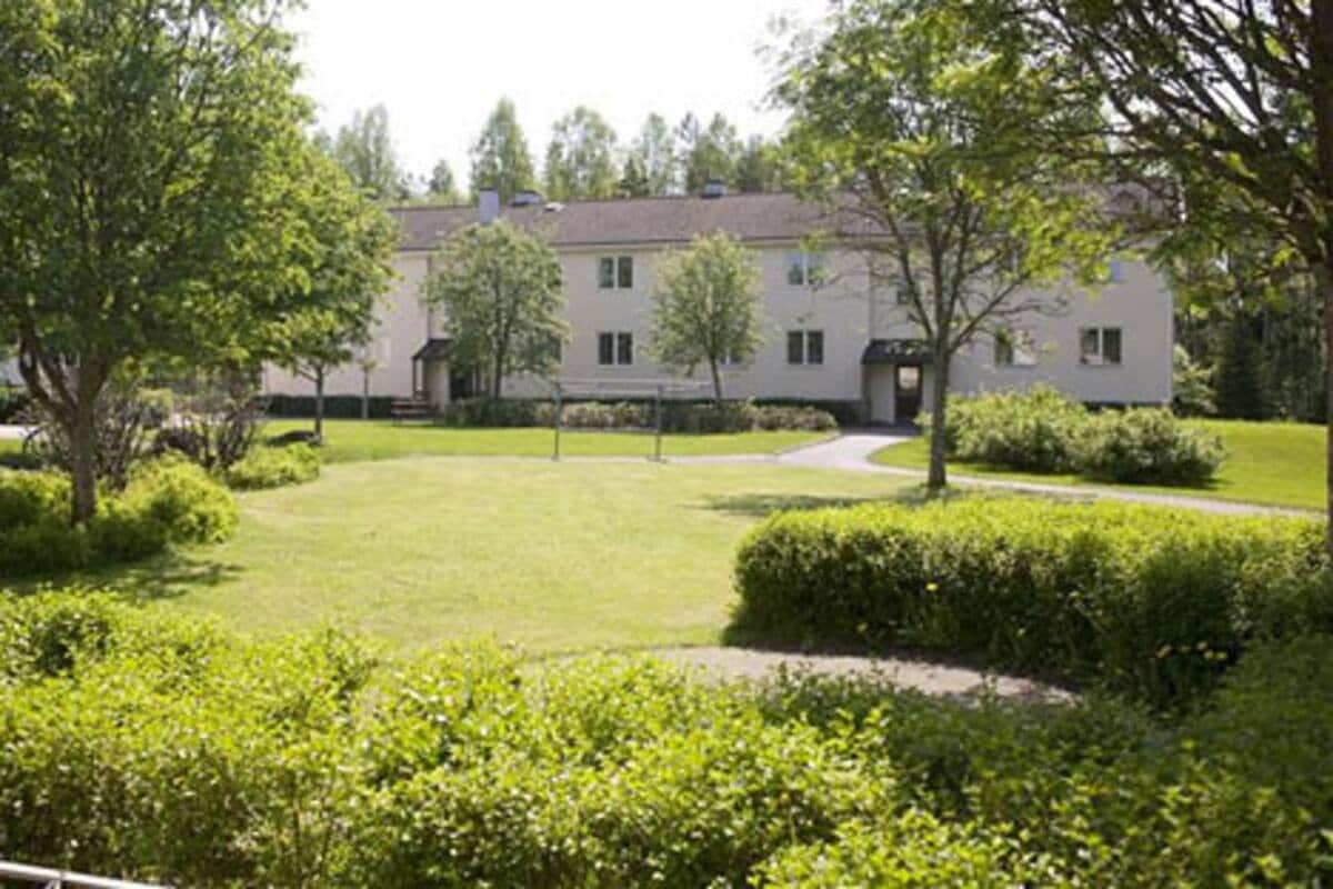 A well-maintained garden area surrounds a two-story building, highlighted by lush green trees and manicured hedges. A pathway meanders through the lawn, leading to the entrance of the building, which features multiple windows and a simple facade.