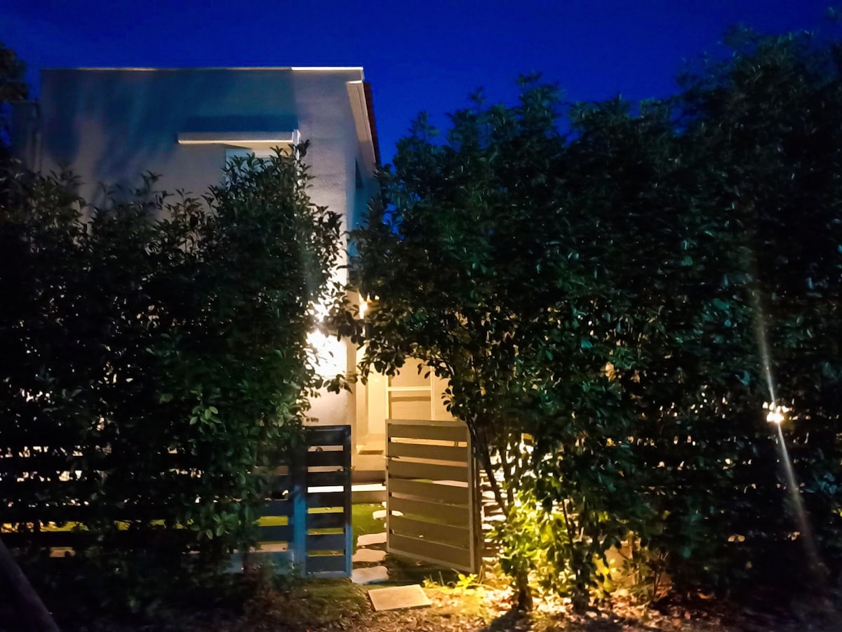 The exterior of the villa is softly illuminated at night, surrounded by lush greenery. A welcoming pathway leads to the door, framed by a wooden fence. Warm lights highlight the entrance, creating a serene and inviting ambiance.