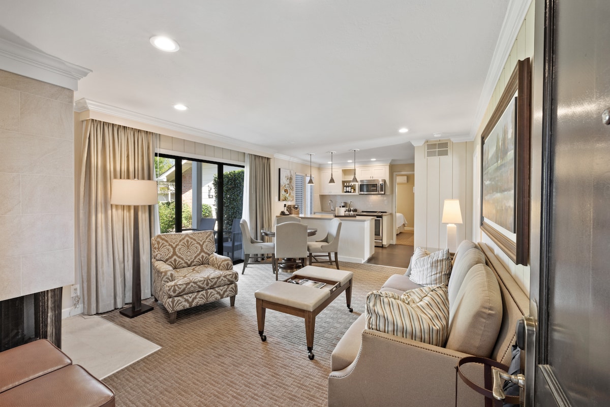 A spacious living area features comfortable seating arrangements, including a cozy sofa and an upholstered chair. Natural light filters through large windows dressed with soft curtains. A dining area and full kitchen are visible in the background, connecting the open layout seamlessly.