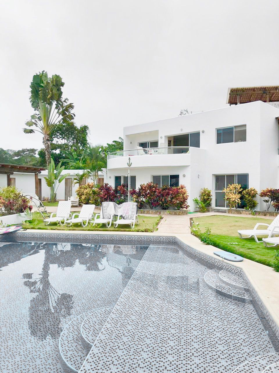 A modern two-story building is set within a landscaped area, featuring a refreshing pool surrounded by lounge chairs. Colorful tropical plants and palm trees enhance the space. A balcony is visible on the second floor, overlooking the inviting pool area.