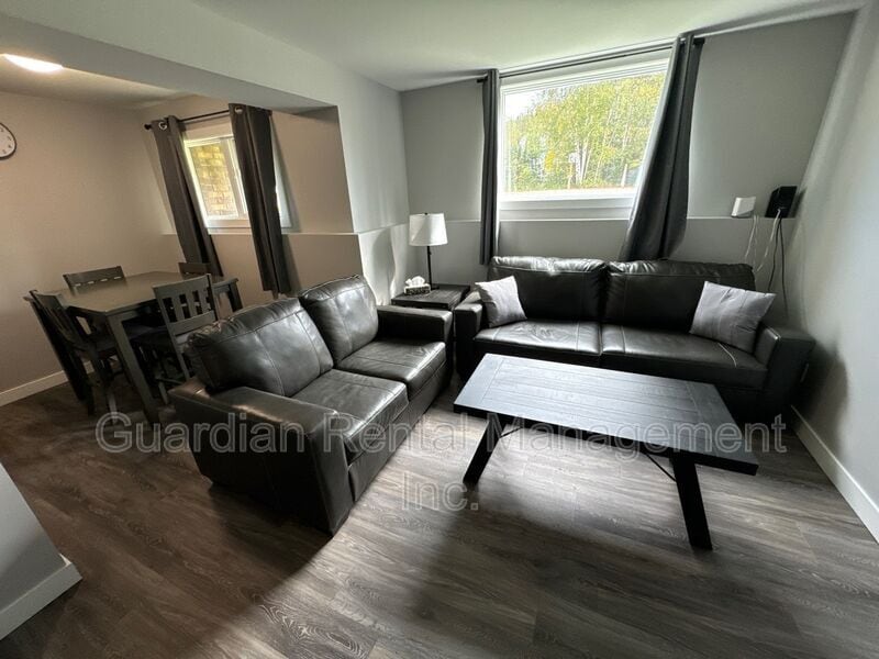 A comfortable living area features a dark leather couch and matching armchair, positioned around a dark coffee table. Natural light enters through a large window, with sheer curtains allowing a soft glow. A dining table is visible in the background, accommodating several chairs.