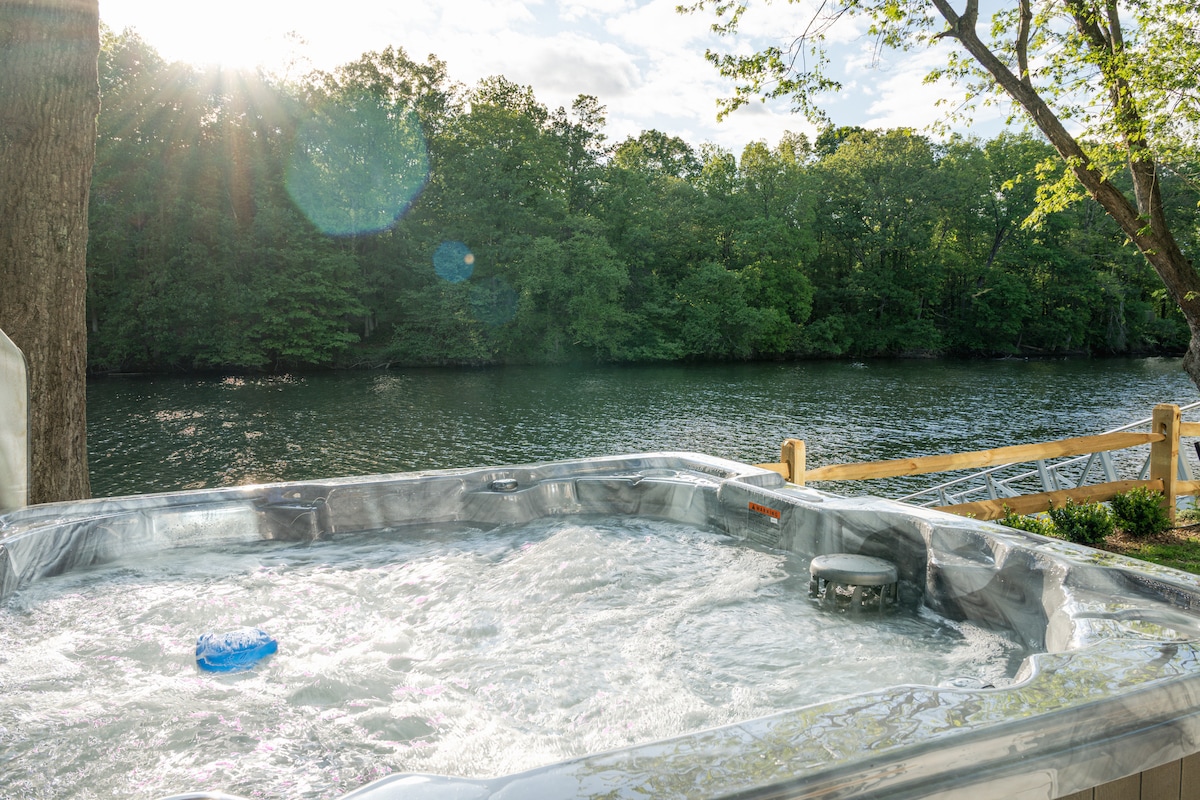 A large hot tub is positioned on a deck overlooking a tranquil river. Bubbles rise from the water's surface, while sunlight filters through the surrounding trees, casting a soft glow over the area. The serene landscape features lush greenery along the water's edge.