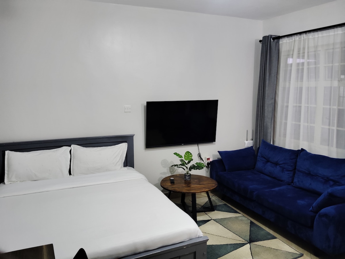 A studio space features a spacious bed with crisp white linens, situated next to a modern blue sofa. A round coffee table is placed nearby, and a flat-screen television is mounted on the wall. Natural light fills the room through a large window with sheer curtains.