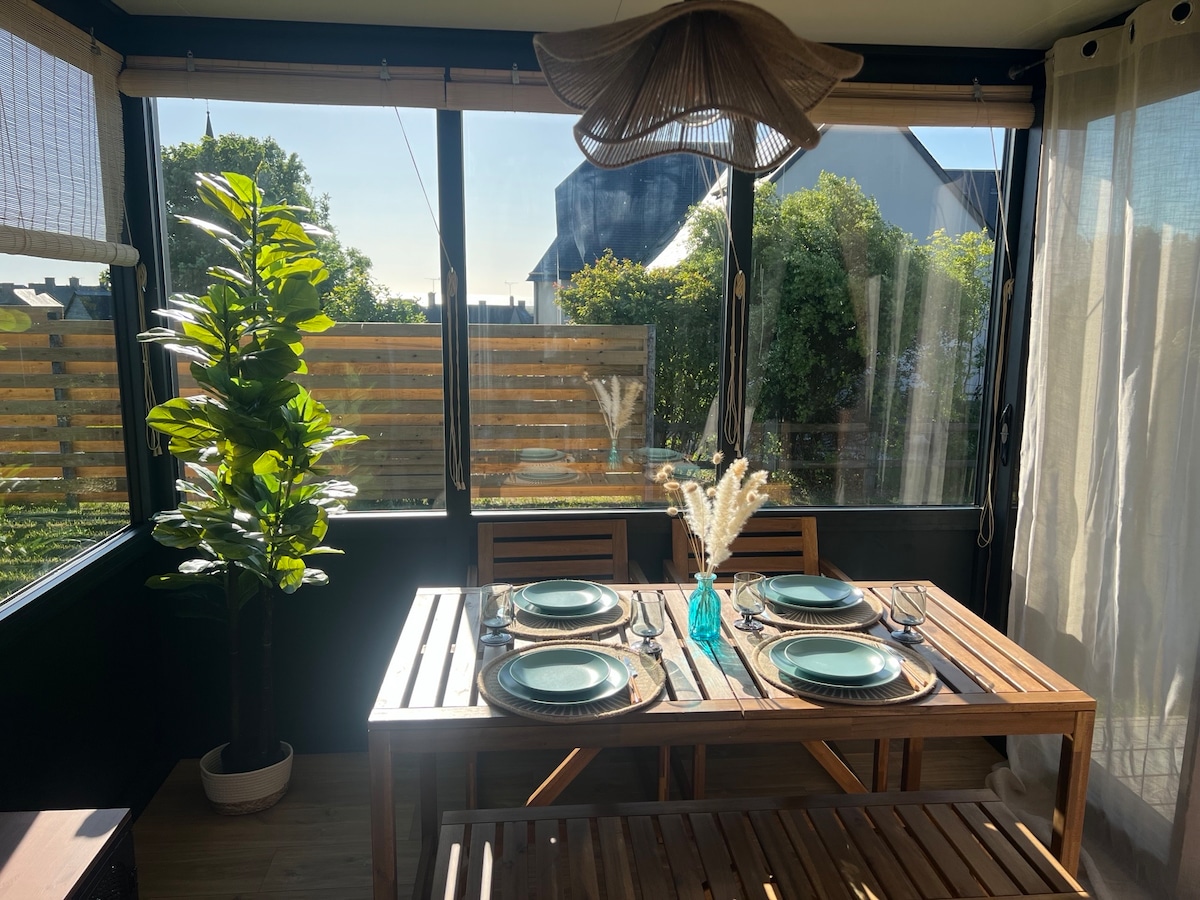 A bright dining area features a wooden table set for four, with turquoise plates and silverware neatly arranged. Large windows provide views of the outdoor greenery, while a potted plant adds a touch of nature to the space. Soft curtains filter the sunlight.