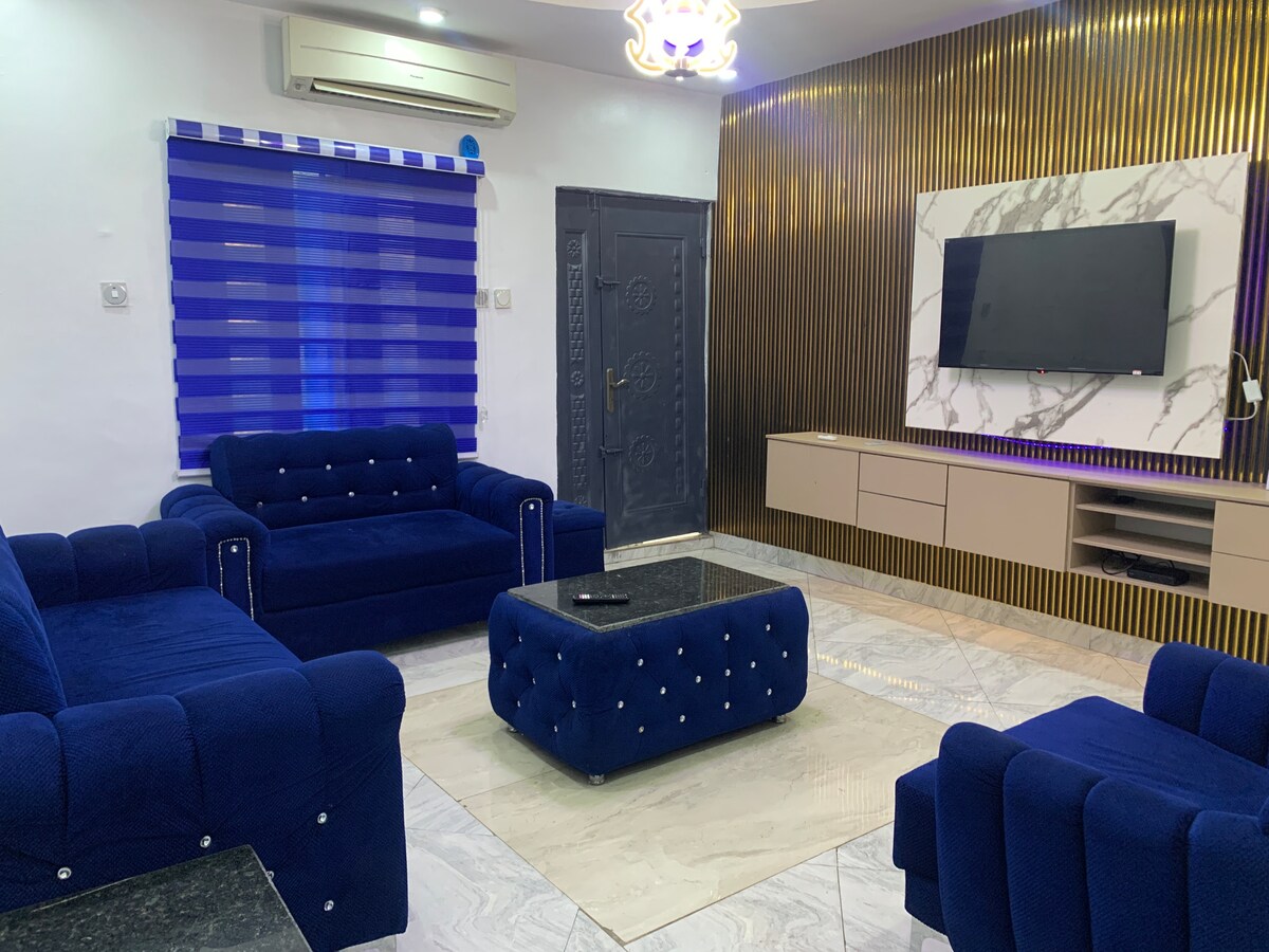 A modern living area is showcased with deep blue upholstered sofas surrounding a sleek, dark marble coffee table. The wall features a stylish design with golden accents, and a large television is mounted for viewing. Natural light filters through a striped window covering.