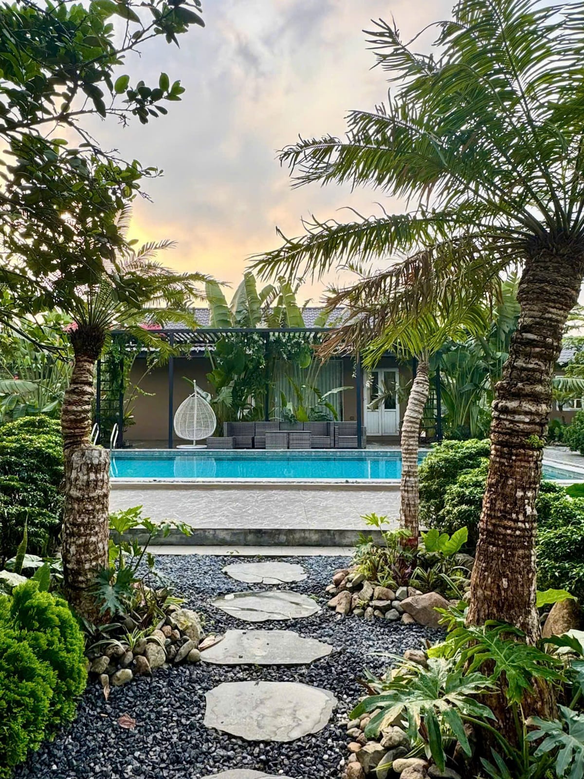 A serene pathway lined with stones leads to a swimming pool, surrounded by lush greenery. Tall palm trees frame the view, while a modern resting area with a hanging chair is visible in the background. Soft light filters through the leaves, creating a tranquil ambiance.