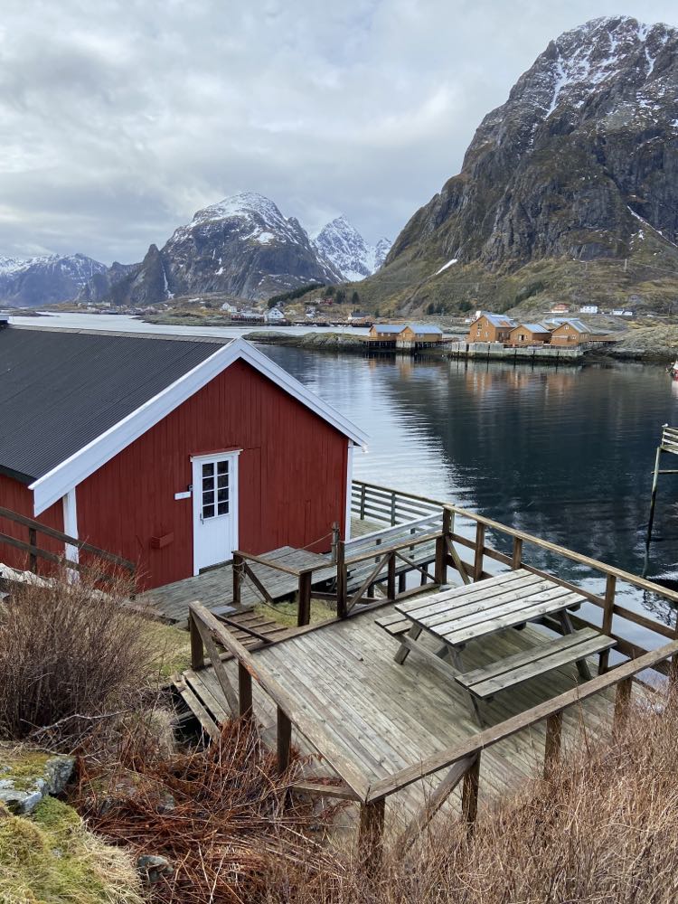 A red rorbu is positioned by the water's edge, featuring a wooden deck with picnic tables. The surrounding landscape includes snow-capped mountains and a calm fjord, creating a serene backdrop for outdoor relaxation.