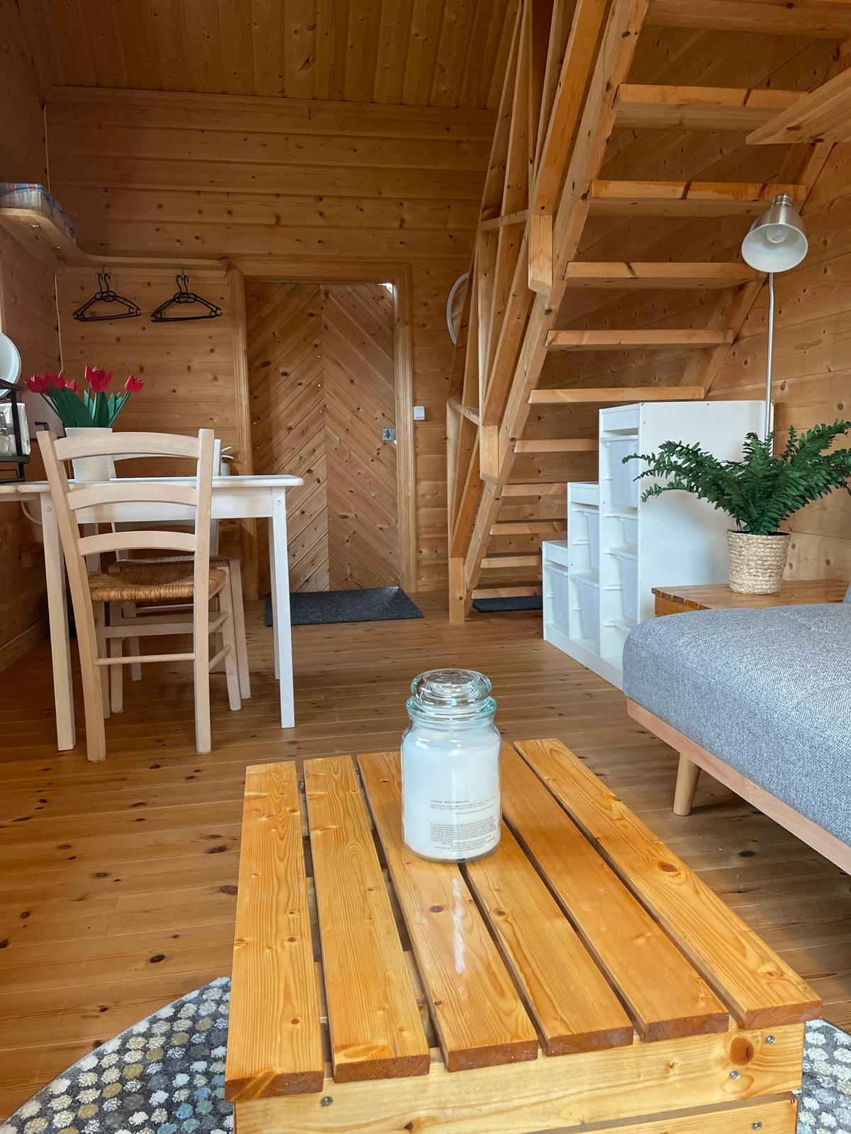 The interior showcases warm wooden tones throughout, featuring a small dining table with chairs and a cozy living area with a sofa. A decorative potted plant adds a touch of greenery. A staircase leads to the upper level, while a wooden crate sits in the foreground.