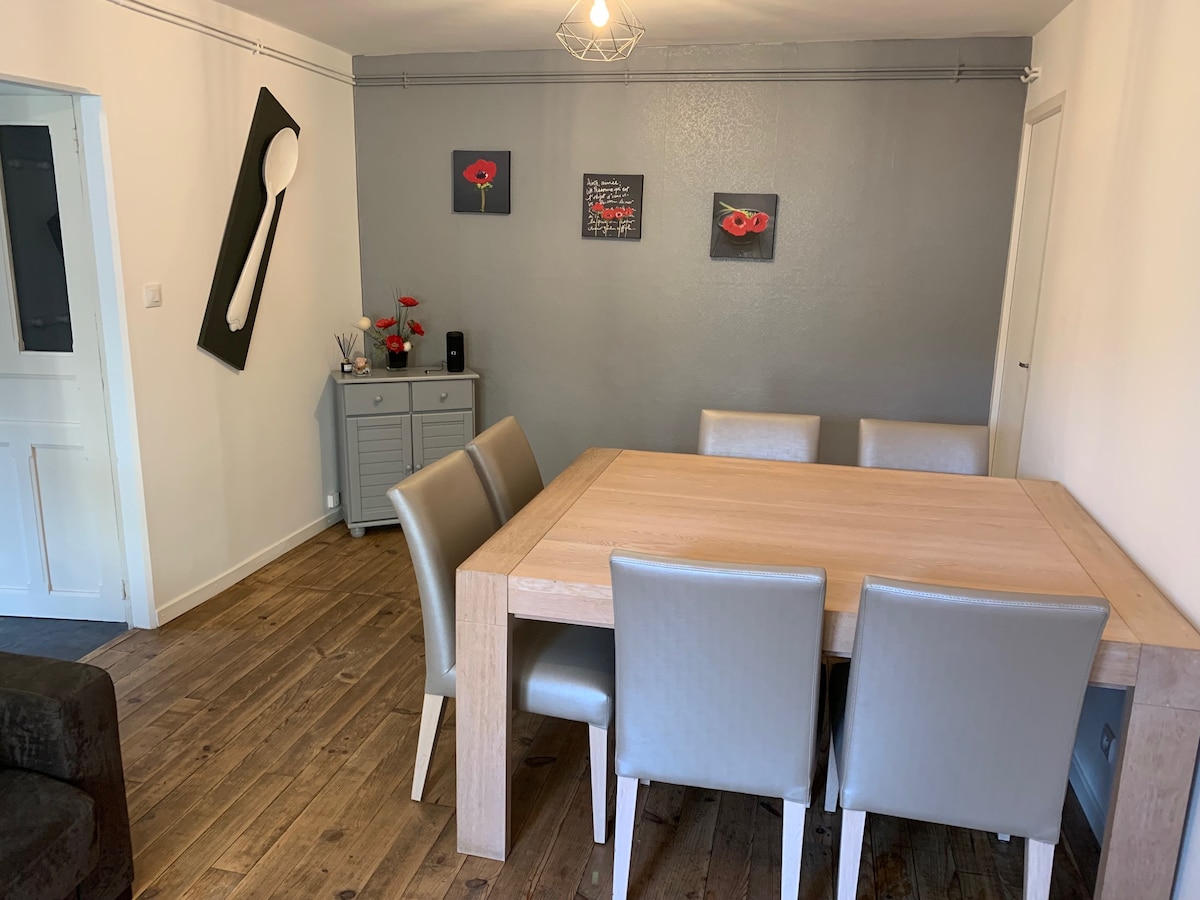 A modern dining area is highlighted by a light wooden table surrounded by six padded chairs. Artistic wall decor featuring floral motifs adds color to the gray walls. A sideboard provides additional storage and complements the minimalist design, while natural light fills the space.