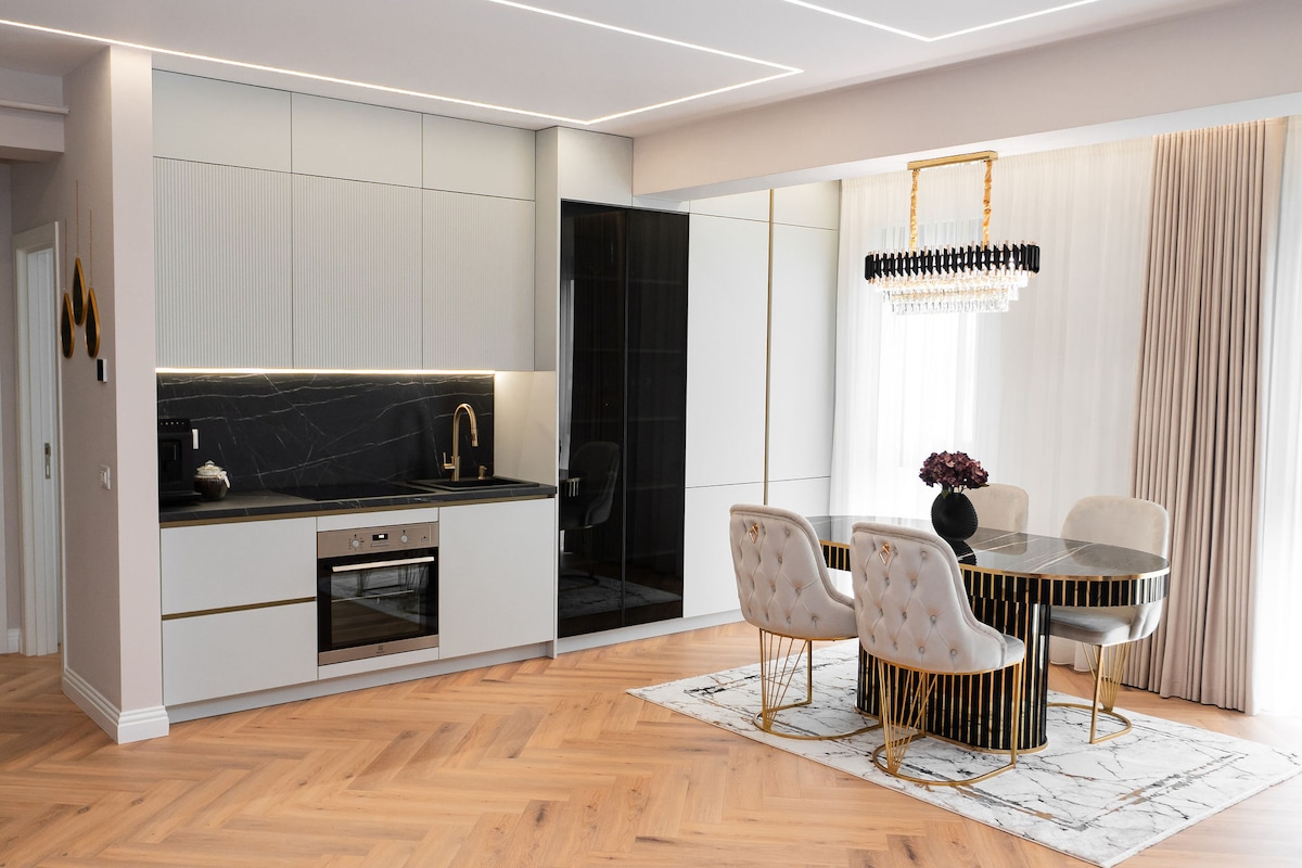 A modern kitchen is displayed, featuring sleek cabinetry in light tones. A black marble countertop contrasts with the white cabinetry. A stylish dining area includes a round table with gold accents and plush chairs. Natural light filters through sheer curtains, enhancing the ambiance.