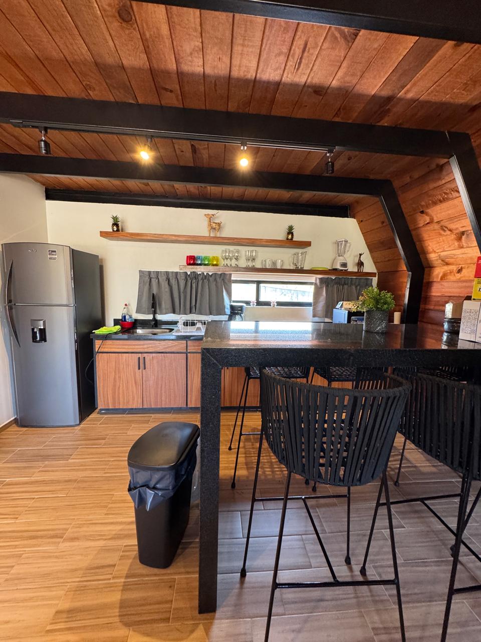 A modern kitchen area features a sleek black countertop with bar stools. Storage shelves hold decorative items, while appliances, including a refrigerator, are positioned along the wall. Natural light illuminates the space through a window, complementing the warm tones of the wooden ceiling.