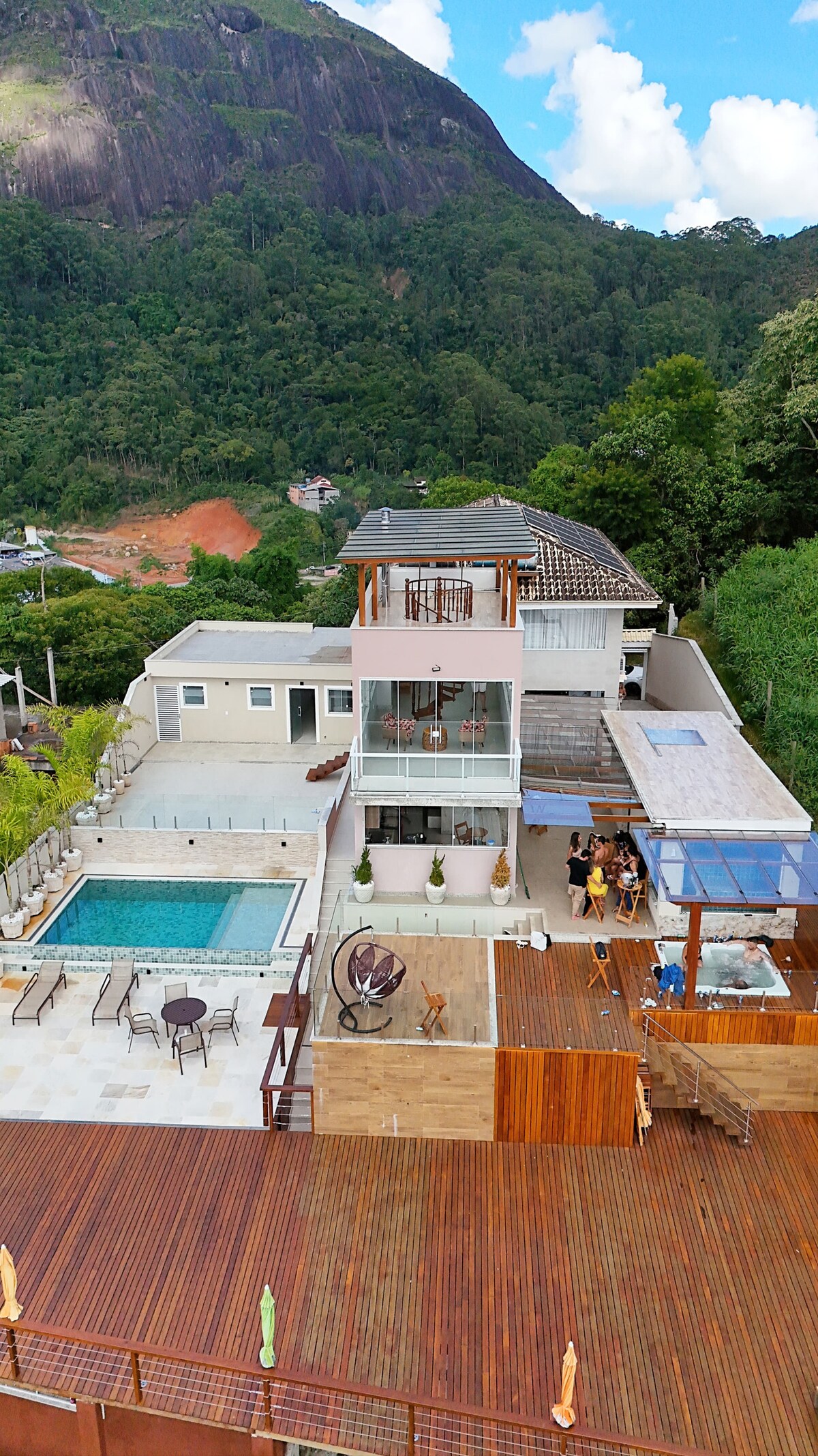 An expansive outdoor area features a large deck with several seating arrangements. A swimming pool is visible, complemented by lush greenery and mountainous terrain in the background. A modern two-story building showcases various architectural details, including large windows and outdoor spaces.