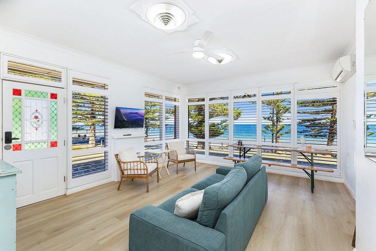 The living area features a comfortable sofa facing large windows that offer views of the beach and ocean. Natural light fills the space, highlighting the wood flooring and decor. A flat-screen television is mounted on the wall, adding to the inviting ambiance of the room.